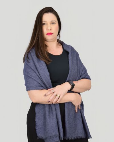 Foto de Alícia Lizz Maia Lopes Pinheiro, colaboradora no escritório de advocacia Gelson Ferrareze. Ela está sorrindo, com cabelos loiros longos soltos, vestindo uma blusa preta de gola alta e um colar dourado com um pingente de cruz. Seus braços estão cruzados, e ela usa um xale azul escuro sobre os ombros. O fundo é neutro e escuro, destacando sua imagem.
